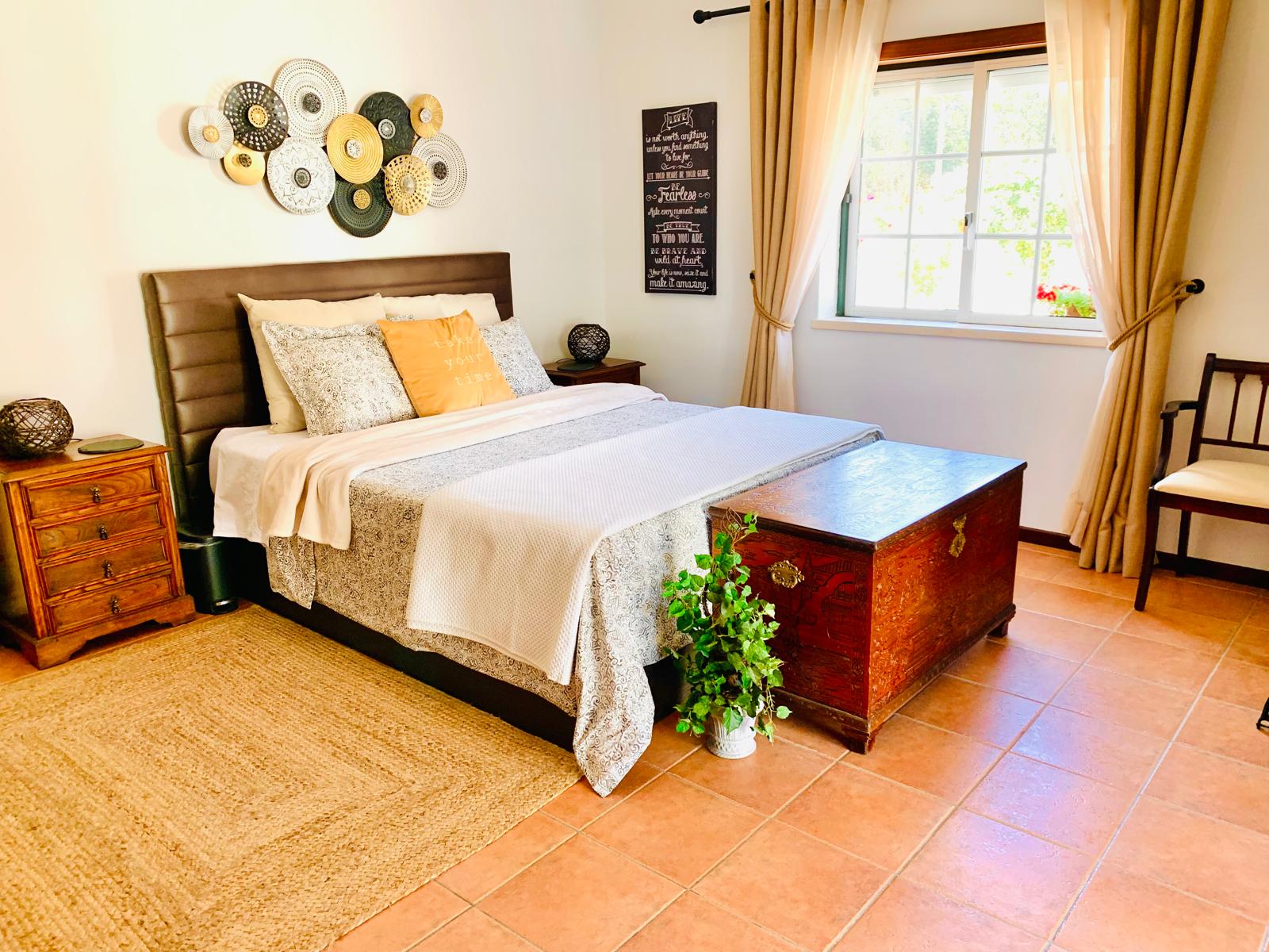 Queen bed with patterned linens, carved wooden chest at the foot and window with garden view